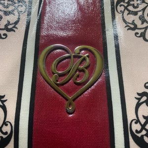 Może przedstawiać: Zbliżenie torebki z pozłacanym, w kształcie serca emblematem z literą "B". Torba ma czerwony panel środkowy, otoczony pionowymi paskami w kolorze czarnym, białym i r&oacute;żowym. Widoczne są dekoracyjne czarne wzory kwiatowe.