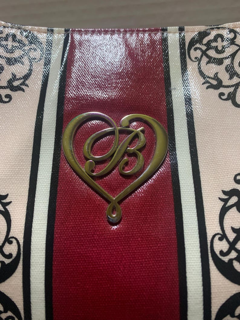 Może przedstawiać: Zbliżenie torebki z pozłacanym, w kształcie serca emblematem z literą "B". Torba ma czerwony panel środkowy, otoczony pionowymi paskami w kolorze czarnym, białym i r&oacute;żowym. Widoczne są dekoracyjne czarne wzory kwiatowe.