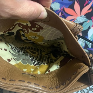 Op de afbeelding: Een bruine leren handtas met een magnetische drukknoopsluiting en zichtbare stiksels langs de randen. De binnenkant is gevoerd met een cr&egrave;mekleurige stof met een herhalend patroon van bruine, gele en groene bloem- en geometrische vormen.