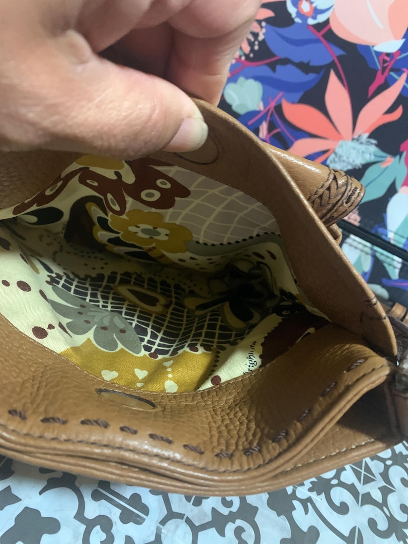Op de afbeelding: Een bruine leren handtas met een magnetische drukknoopsluiting en zichtbare stiksels langs de randen. De binnenkant is gevoerd met een cr&egrave;mekleurige stof met een herhalend patroon van bruine, gele en groene bloem- en geometrische vormen.
