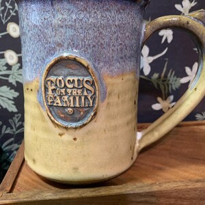 May include: A ceramic mug with a two-tone glaze featuring a light blue top and a tan bottom. An oval medallion is centered on the front with the embossed text "FOCUS ON THE FAMILY" and a registered trademark symbol. The mug has a sturdy handle.