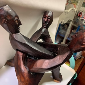 May include: A dark brown wooden sculpture featuring three stylized human figures linked together in a circular formation. Each figure has a smooth, carved face and elongated body, with their arms interlocked to form a continuous, connected structure.