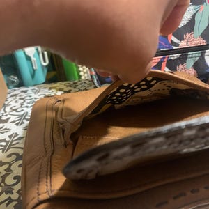 Op de afbeelding: Een bruine leren handtas met een getextureerd oppervlak en zichtbare stiksels langs de randen. De tas heeft een open flap die een binnenvoering onthult met een abstract patroon in cr&egrave;me, zwart en geel. De buitenkant heeft een gestructureerd ontwerp.