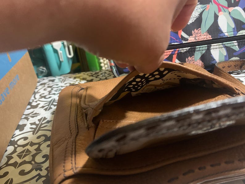Op de afbeelding: Een bruine leren handtas met een getextureerd oppervlak en zichtbare stiksels langs de randen. De tas heeft een open flap die een binnenvoering onthult met een abstract patroon in cr&egrave;me, zwart en geel. De buitenkant heeft een gestructureerd ontwerp.