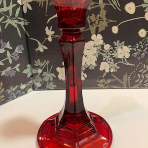 May include: A tall, ruby red glass candlestick holder featuring a faceted, geometric stem and a wide, circular base. The top section has a flared rim designed to hold a taper candle, with the entire piece crafted from translucent, deep red glass.