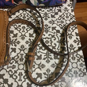 Op de afbeelding: Een beige lederen schouderband voor een tas met decoratieve stiksels langs de randen en een zilverkleurige metalen gesp voor lengteverstelling. De band is verbonden met een beige lederen tas met zichtbare stiksels en een zilveren ring.
