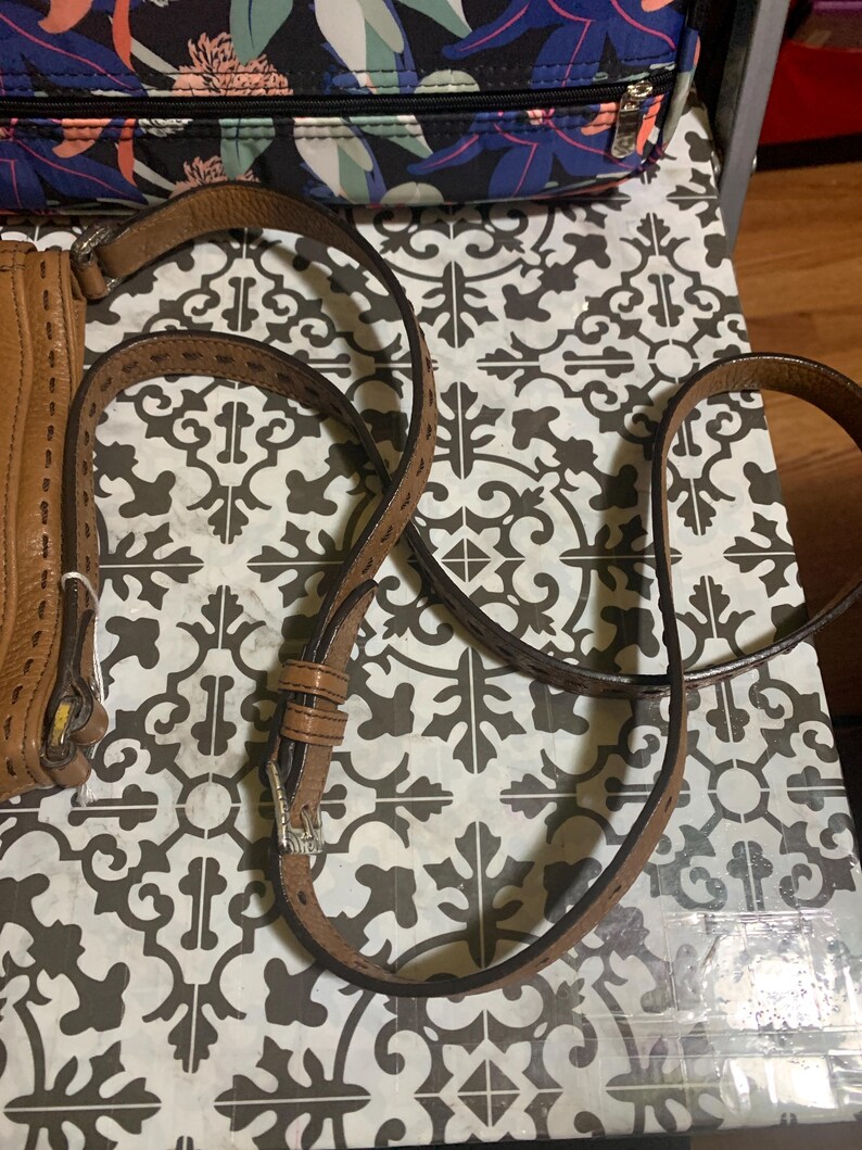 Op de afbeelding: Een beige lederen schouderband voor een tas met decoratieve stiksels langs de randen en een zilverkleurige metalen gesp voor lengteverstelling. De band is verbonden met een beige lederen tas met zichtbare stiksels en een zilveren ring.