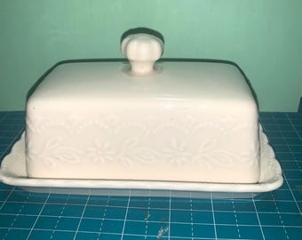 May include: Off-white ceramic butter dish with a lid and a decorative floral pattern. The dish has a rectangular shape with a scalloped edge on the base and a small knob on the lid. The butter dish is designed for kitchen use.