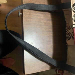 Może przedstawiać: Czarny uchwyt torby typu tote łukuje się nad drewnianą powierzchnią. Wz&oacute;r materiału torby jest widoczny po prawej stronie. Uchwyt wykonany jest z mocnego, tkanego materiału. Drewniana powierzchnia ma ciepły, brązowy odcień.