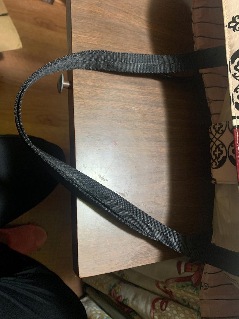 Może przedstawiać: Czarny uchwyt torby typu tote łukuje się nad drewnianą powierzchnią. Wz&oacute;r materiału torby jest widoczny po prawej stronie. Uchwyt wykonany jest z mocnego, tkanego materiału. Drewniana powierzchnia ma ciepły, brązowy odcień.