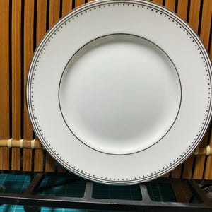 May include: A white ceramic plate with a black dotted border. The plate is round and has a slightly raised rim.