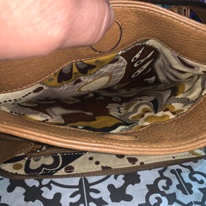 Op de afbeelding: Een bruine leren handtas met een ritssluiting aan de bovenkant. De binnenkant is gevoerd met een cr&egrave;mekleurige stof met een bruin, geel en wit bloemen- en paisleypatroon. De tas heeft een buitenvak met rits met dezelfde voering.