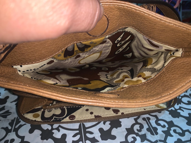 Op de afbeelding: Een bruine leren handtas met een ritssluiting aan de bovenkant. De binnenkant is gevoerd met een cr&egrave;mekleurige stof met een bruin, geel en wit bloemen- en paisleypatroon. De tas heeft een buitenvak met rits met dezelfde voering.