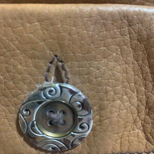 Op de afbeelding: Een bruin, getextureerd leren oppervlak met een ronde knoop met een metallic zilveren afwerking en gegraveerde krulpatronen. De knoop heeft een centraal ontwerp met vier gaten en is met donker garen bevestigd aan het gevlochten leer.