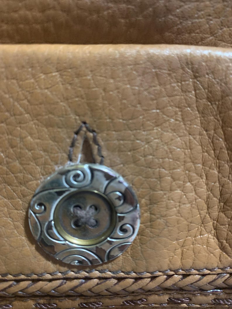 Op de afbeelding: Een bruin, getextureerd leren oppervlak met een ronde knoop met een metallic zilveren afwerking en gegraveerde krulpatronen. De knoop heeft een centraal ontwerp met vier gaten en is met donker garen bevestigd aan het gevlochten leer.