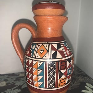 May include: A terracotta pitcher with a matching lid and a single handle. The body features a band of geometric patterns in black, white, orange, and teal, including triangles, diamonds, and cross-hatched lines against the natural clay surface.