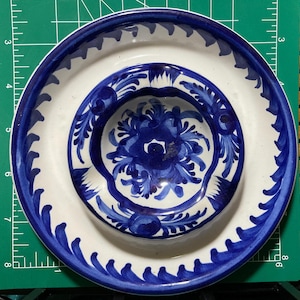 May include: A set of three ceramic plates with a white base and blue floral and leaf patterns. The largest plate has a scalloped edge, and the smallest is a small bowl. The plates are nested on a green cutting mat.