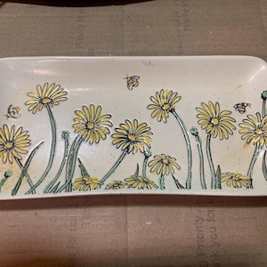 May include: A rectangular off-white ceramic tray featuring a speckled finish and a hand-painted design of yellow daisies with green stems and leaves. Small flying bees are interspersed among the flowers across the surface of the tray.
