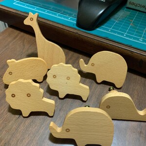 May include: A collection of wooden animal figurines, including a giraffe, hippopotamus, lion, and elephant. The figurines are light brown and have simple, rounded designs. The giraffe stands tall, while the other animals are more compact.