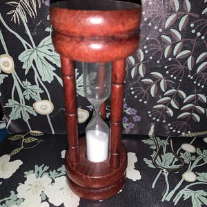 May include: A wooden hourglass with a dark brown finish, featuring three vertical support pillars connecting a circular top and base. The central glass chamber contains white sand, with the sand currently settled in the bottom bulb.