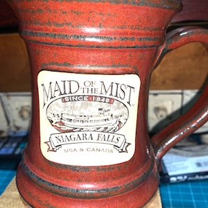 May include: A red ceramic mug with a handle, featuring a "Maid of the Mist" label with an illustration of a boat at Niagara Falls. The mug has a rustic, textured finish and the text "USA & CANADA" is visible.