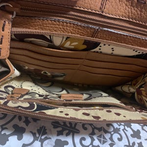 Op de afbeelding: De binnenkant van een bruine leren handtas met een ritsvak, meerdere kaartsleuven en een cr&egrave;mekleurige voering met een bruin, geel en zwart bloemenpatroon. De leren randen zijn afgewerkt met zichtbare bruine stiksels.