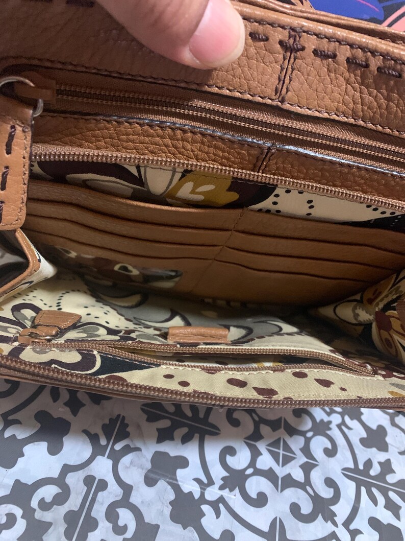 Op de afbeelding: De binnenkant van een bruine leren handtas met een ritsvak, meerdere kaartsleuven en een cr&egrave;mekleurige voering met een bruin, geel en zwart bloemenpatroon. De leren randen zijn afgewerkt met zichtbare bruine stiksels.