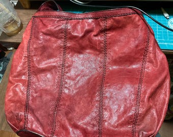 May include: A red leather shoulder bag with a rounded shape. The bag features vertical stitched detailing and a handle at the top. The leather has a distressed appearance, giving it a vintage look.