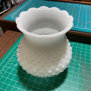 May include: A white hobnail vase with a scalloped rim. The vase has a rounded body covered in small, raised bumps. The vase is empty and sits on a green cutting mat.
