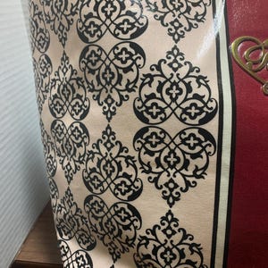 Może przedstawiać: Torba typu tote z kremowym tłem i powtarzającym się czarnym wzorem ornamentalnym. Torba ma czerwony panel ze złotym elementem dekoracyjnym. Czarno-biały pasek oddziela wzorzystą część od czerwonego panelu.