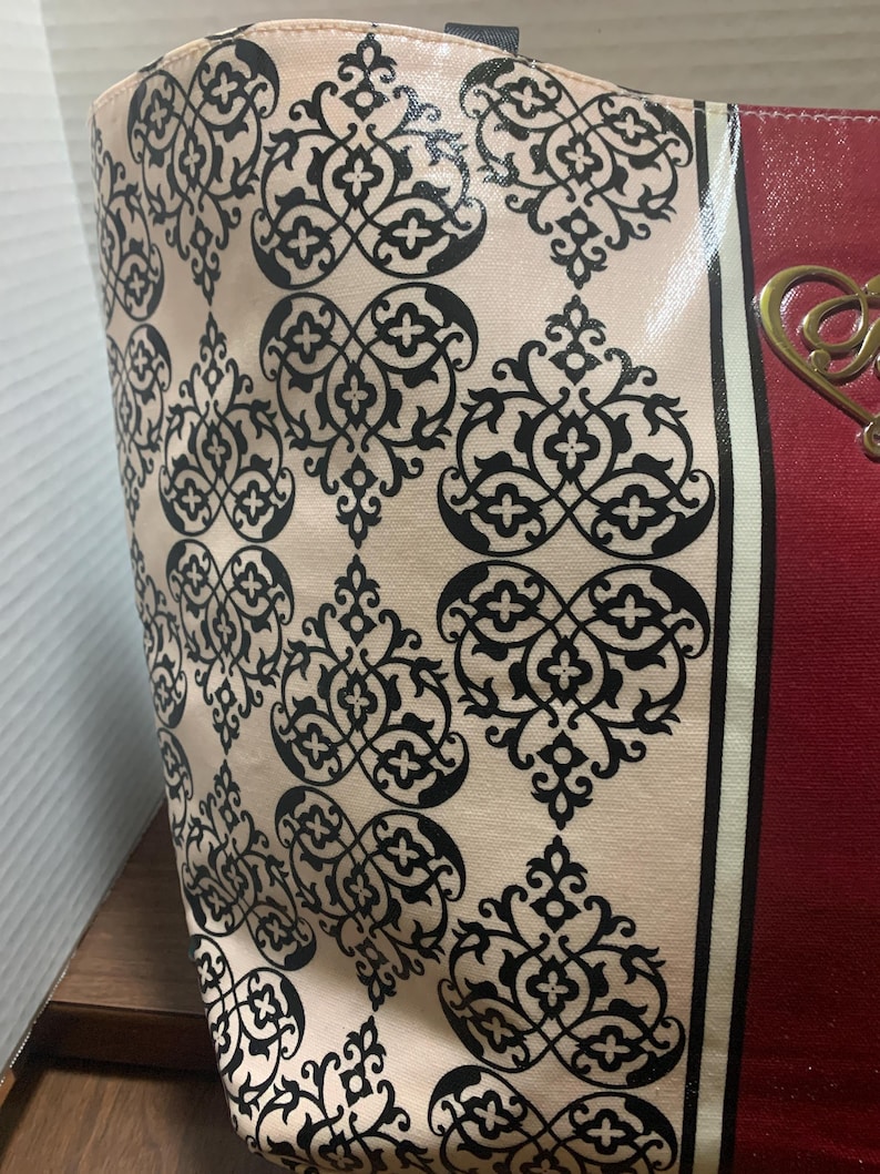 Może przedstawiać: Torba typu tote z kremowym tłem i powtarzającym się czarnym wzorem ornamentalnym. Torba ma czerwony panel ze złotym elementem dekoracyjnym. Czarno-biały pasek oddziela wzorzystą część od czerwonego panelu.