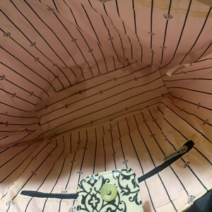 Może przedstawiać: Widok od wewnątrz torby typu tote z r&oacute;żową i czarną podszewką w paski. Podszewka ma powtarzający się wz&oacute;r małych emblemat&oacute;w. Widoczna jest mała, wzorzysta klapka z zapięciem na guzik. Widoczny jest r&oacute;wnież czarny zamek błyskawiczny.