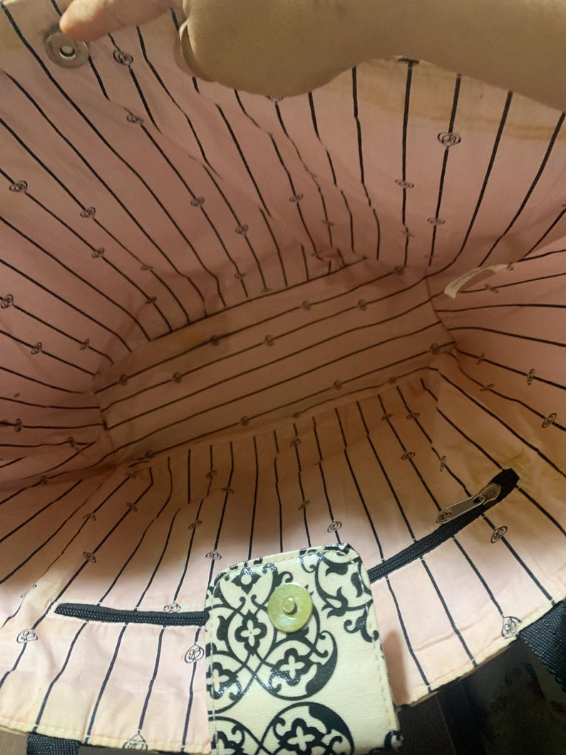 Może przedstawiać: Widok od wewnątrz torby typu tote z r&oacute;żową i czarną podszewką w paski. Podszewka ma powtarzający się wz&oacute;r małych emblemat&oacute;w. Widoczna jest mała, wzorzysta klapka z zapięciem na guzik. Widoczny jest r&oacute;wnież czarny zamek błyskawiczny.