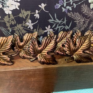 May include: A set of six metallic napkin rings shaped like maple leaves, featuring a burnished bronze and copper finish with detailed veining. The leaves are arranged in a row, showcasing a warm, reflective surface with dark, recessed accents.