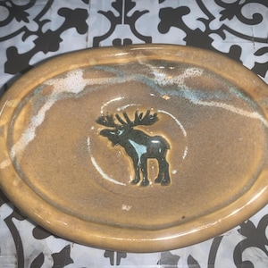 May include: An oval-shaped ceramic soap dish with a glossy tan glaze. The center features a dark brown embossed moose silhouette with a light blue accent on its body. A white, textured glaze border runs along the upper rim of the dish.