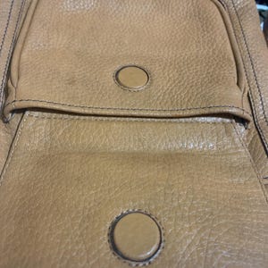 Op de afbeelding: Een tas van getextureerd bruin leer met een voorvak met klep en een ronde magnetische sluiting. De tas heeft zichtbare donkere stiksels langs de randen en een tweede bijpassende ronde magnetische drukknoop op het hoofdgedeelte van de tas.