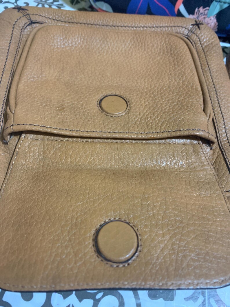 Op de afbeelding: Een tas van getextureerd bruin leer met een voorvak met klep en een ronde magnetische sluiting. De tas heeft zichtbare donkere stiksels langs de randen en een tweede bijpassende ronde magnetische drukknoop op het hoofdgedeelte van de tas.