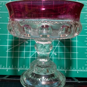 May include: A clear glass pedestal bowl with a deep red rim. The bowl has a scalloped edge and a textured design.
