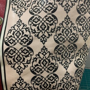 Może przedstawiać: Zbliżenie torby typu tote z kremowym tłem i powtarzającym się czarnym wzorem kwiatowym. Torba ma czerwony pasek z boku ze złotym oczkiem. Torba wykonana jest z błyszczącego, trwałego materiału.