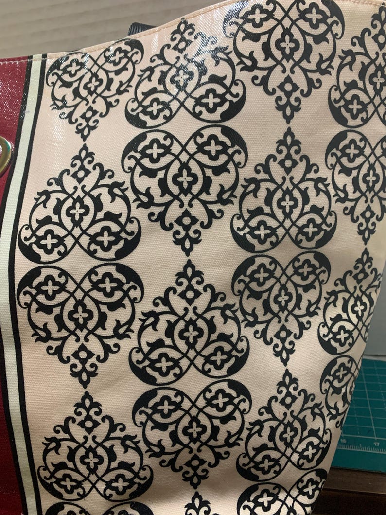 Może przedstawiać: Zbliżenie torby typu tote z kremowym tłem i powtarzającym się czarnym wzorem kwiatowym. Torba ma czerwony pasek z boku ze złotym oczkiem. Torba wykonana jest z błyszczącego, trwałego materiału.