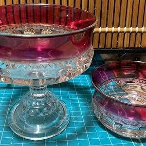 May include: Two clear glass bowls with a red and clear iridescent design. The larger bowl is on a pedestal and the smaller bowl is sitting on a blue and green surface.