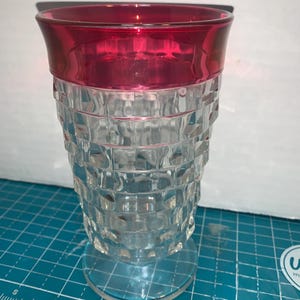 May include: A clear glass goblet with a red rim. The body of the glass has a textured, square pattern. The goblet sits on a clear, circular base. The glass is likely used for beverages.