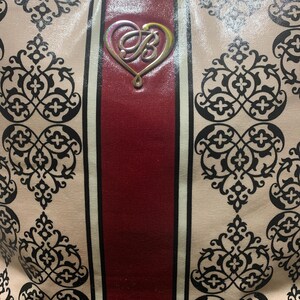 Może przedstawiać: Beżowa torba typu tote z powtarzającym się czarnym wzorem kwiatowym. Torba ma pionowy bordowy pasek pośrodku, otoczony białymi paskami. Srebrny emblemat w kształcie serca ze stylizowanym "B" jest wyśrodkowany na bordowym pasku.