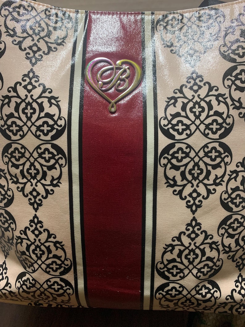 Może przedstawiać: Beżowa torba typu tote z powtarzającym się czarnym wzorem kwiatowym. Torba ma pionowy bordowy pasek pośrodku, otoczony białymi paskami. Srebrny emblemat w kształcie serca ze stylizowanym "B" jest wyśrodkowany na bordowym pasku.