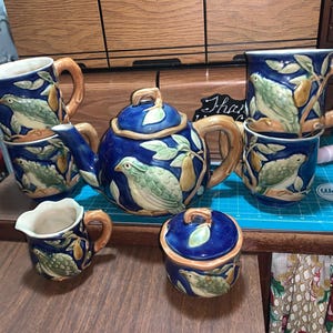 May include: A blue ceramic tea set with a bird and pear design. The set includes a teapot, sugar bowl, creamer, and two stacked mugs. The handles and rims are brown. The set is on a wooden surface.