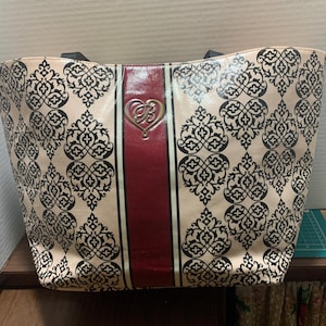 Może przedstawiać: Beżowa torba typu tote z czarnym wzorem w damaszek. Torba posiada pionowy bordowy pas pośrodku, ze złotym emblematem w kształcie serca. Czarne uchwyty dopełniają design.