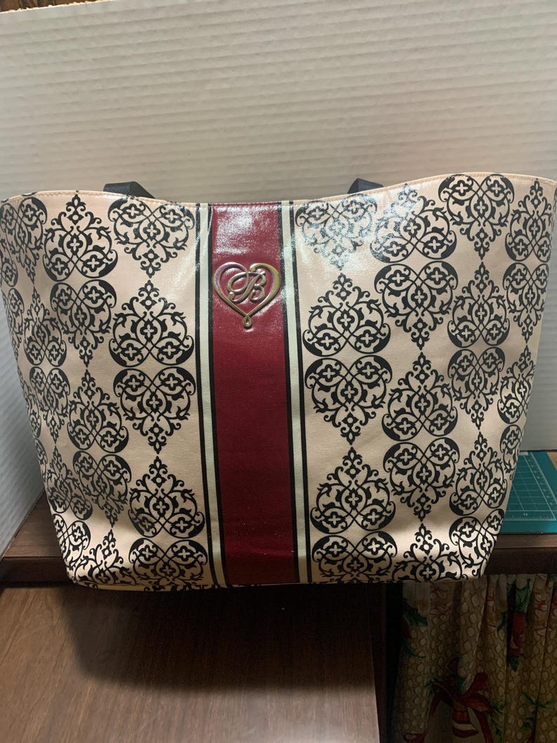 Może przedstawiać: Beżowa torba typu tote z czarnym wzorem w damaszek. Torba posiada pionowy bordowy pas pośrodku, ze złotym emblematem w kształcie serca. Czarne uchwyty dopełniają design.