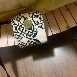 Może przedstawiać: Kremowa torba z czarno-białym kwiatowym wzorem na klapie. Torba ma magnetyczne zapięcie i czarny pasek. Wnętrze torby jest jasnor&oacute;żowe z czarnymi pionowymi paskami.