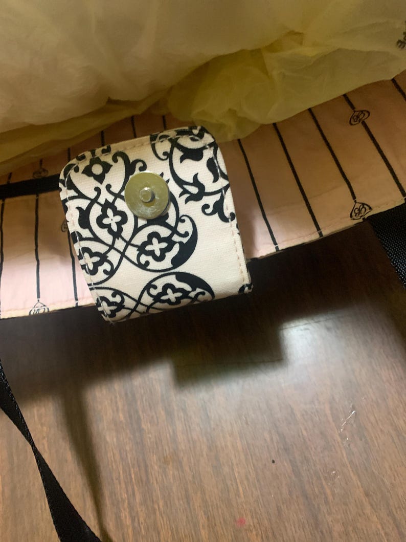 Może przedstawiać: Kremowa torba z czarno-białym kwiatowym wzorem na klapie. Torba ma magnetyczne zapięcie i czarny pasek. Wnętrze torby jest jasnor&oacute;żowe z czarnymi pionowymi paskami.