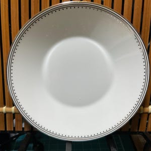 May include: A white ceramic plate with a black dotted border. The plate has a wide rim and a slightly recessed center. The plate is round and appears to be new. The plate is on a wooden surface.